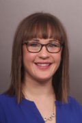 Headshot of a woman with shoulder-length brown hair and bangs, wearing black square-framed glasses and a royal-blue V-neck top.