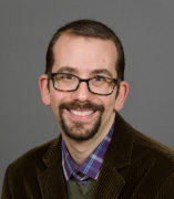 Brian Grossman, a white smiling man with glasses in his mid 30s, dressed in a purple plaid shirt, green sweater, and brown corduroy blazer.