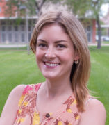 Caitlin is wearing a sleeveless dress with flowers on it, standing behind the DHSP building with grass in the background, with her arms crossed and smiling.