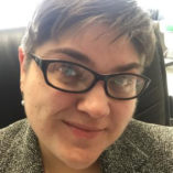 Photograph of a white woman with short brown hair and black glasses seated in her office at DHD
