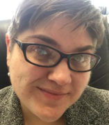 Photograph of a white woman with short brown hair and black glasses seated in her office at DHD