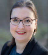 Woman with brown hair, brown eyes and glasses in front of a blurry dark background.