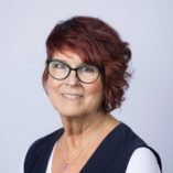 White middle aged female with short red hair wearing dark rimmed glasses and a navy blue and white top.