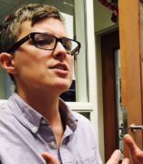 Image of a white woman with short brown hair and dark glasses talking, wearing a light purple dress shirt.