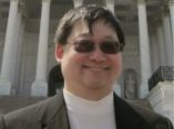 Asian man with dark brown hair, wearing shaded glasses, white shirt, dark grey sport coat.  Behind are white pillars.