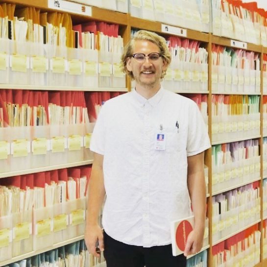 PhD student Drew standing in front of bookshelves of documents