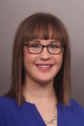 Headshot of a woman with shoulder-length brown hair and bangs, wearing black square-framed glasses and a royal-blue V-neck top.