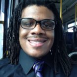 Smiling African-American cis-male person, with locs, black shirt, purple tie, and glasses