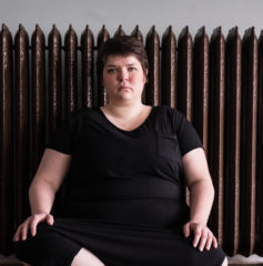 Maggie, a fat, white woman with short brown hair, sits in a dance studio in front of a brown, ornate radiator. Her legs are crossed with her hands on her knees.  