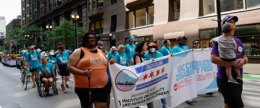 A group of differently abled people affiliated with UIC and its Department of Disability and Human Development march in the 2023 Chicago Disability Pride Parade. At the front of the group there are a number of people holding two horizontal banners.