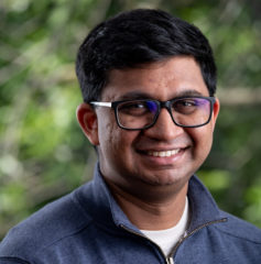 Javin, an Indian-American person with black hair and black glasses, smiles directly at the camera. They are wearing a dark-blue UIC-themed quarter zip jacket and a white shirt. The blurred background includes tree branches and green leaves.