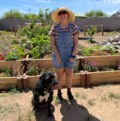 Bri stands against a backdrop of a garden full of greenery wearing a sunhat, sunglasses and overalls. Next to them is their dog, Fable, who is a black lab with white patches on her chest.