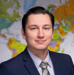 Background of global map, foreground is of Kevin Hans Waitkuweit in a navy suit, light grey shirt, and multicolored tie. Waitkuweit has brown slicked back hair with a side part and a pale complexion.