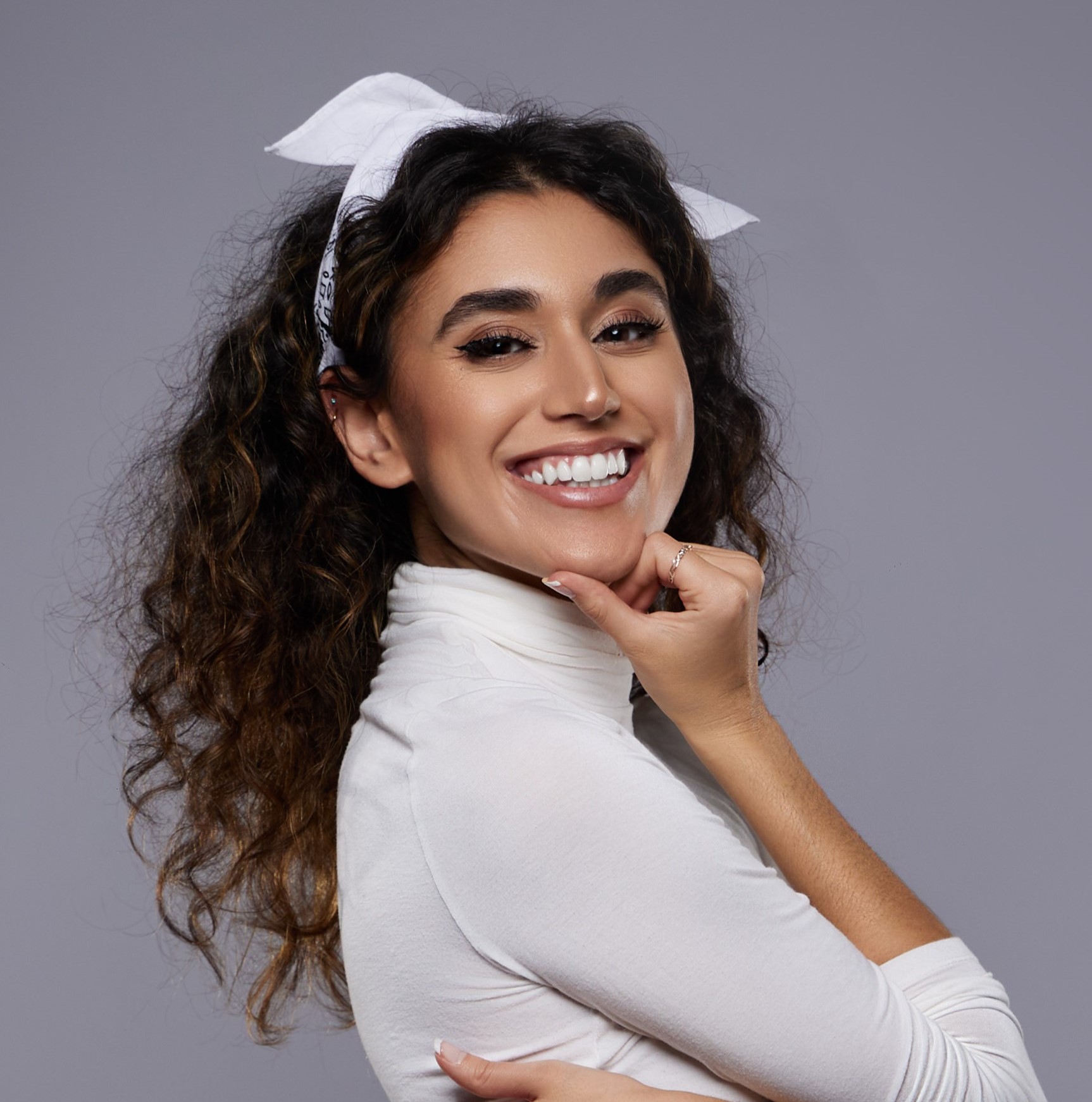 A brown skinned Arab woman with brown eyes and dark brown natural curls. Sabrina is wearing a white turtleneck and white headband. Sabrina wears a big smile on her face.