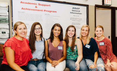 A group of six women sit in front of a bulletin board with ASAP information