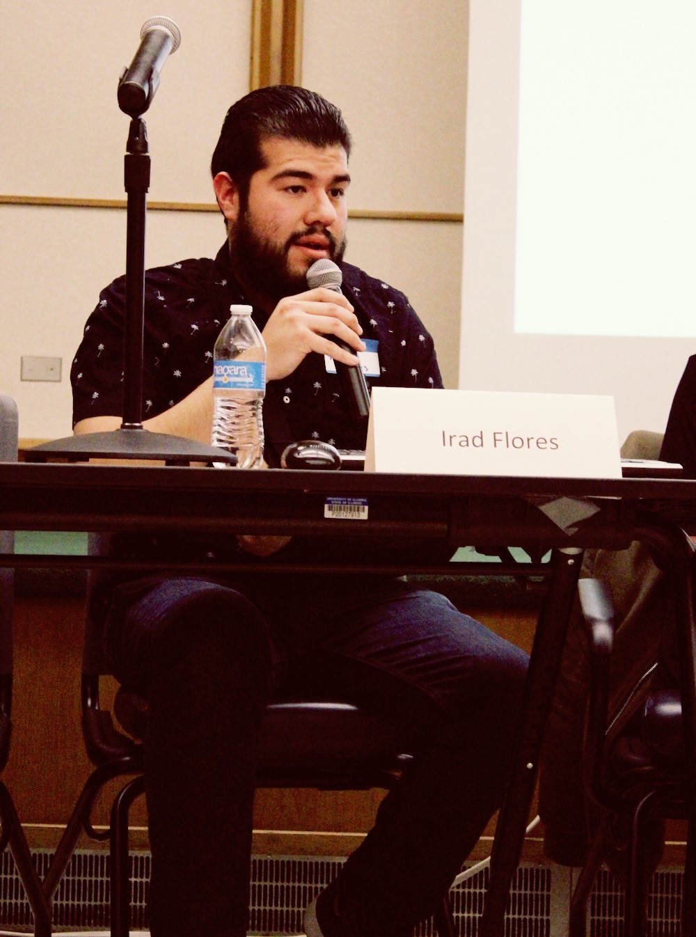 Irad Flores sits behind table holding a microphone near his mouth as he is speaking