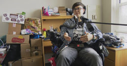 Noah Ohashi sits in a wheelchair in a college dorm room with a microphone in front of them